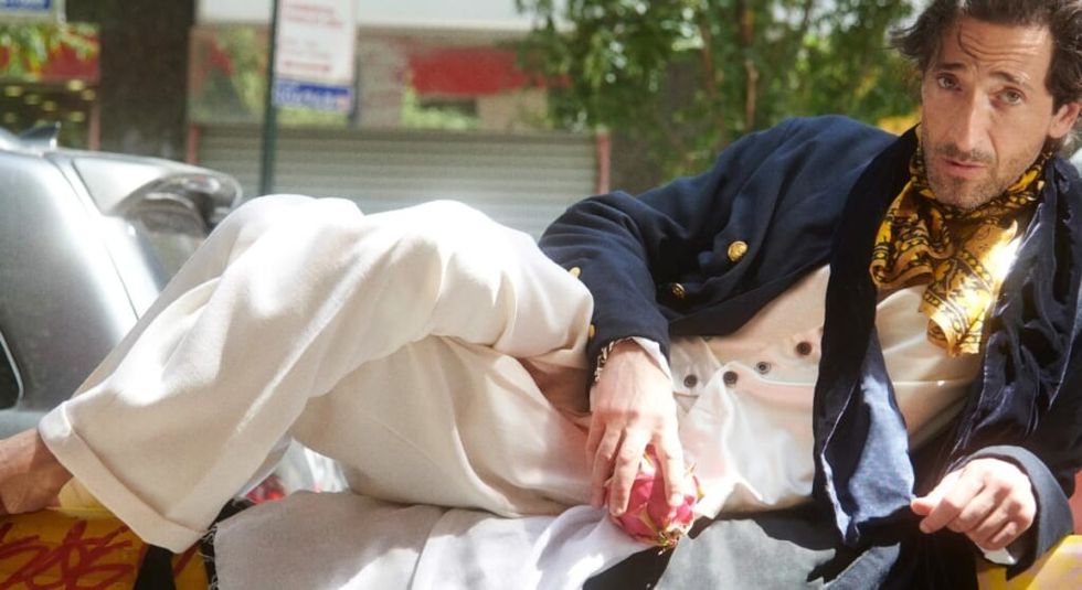 Adrien Brody in navy jacket reclining on a couch and holding an apple.