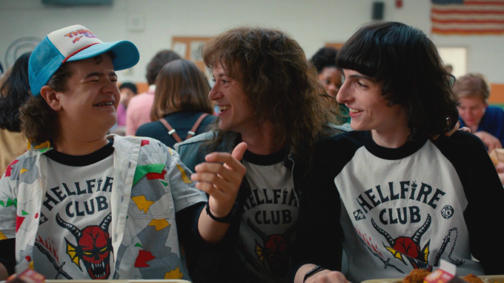 Dustin Henderson, wearing a baseball cap, smiling at Eddie Munson who smiles back at him. On Eddie's left is Mike Wheeler. All three are wearing Hellfire Club shirts.