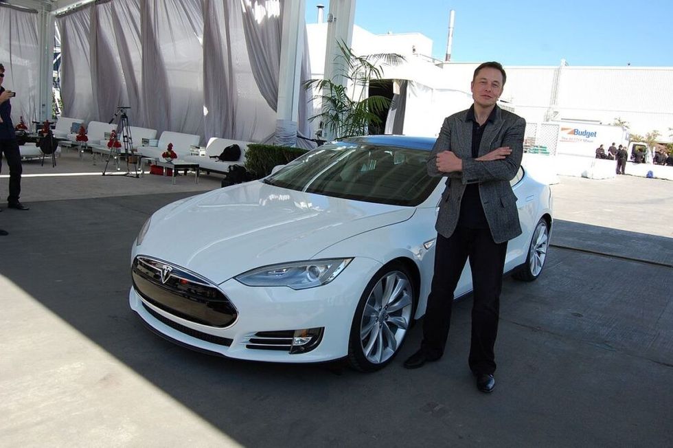 Elon Musk standing in front of white Tesla with arms folded.