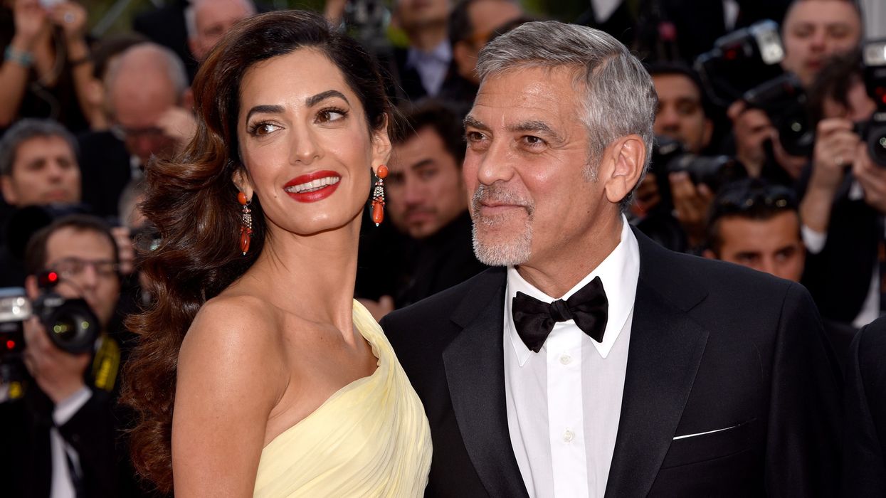 George Clooney in a tux posing with his wife Amal Clooney who is wearing a yellow dress.