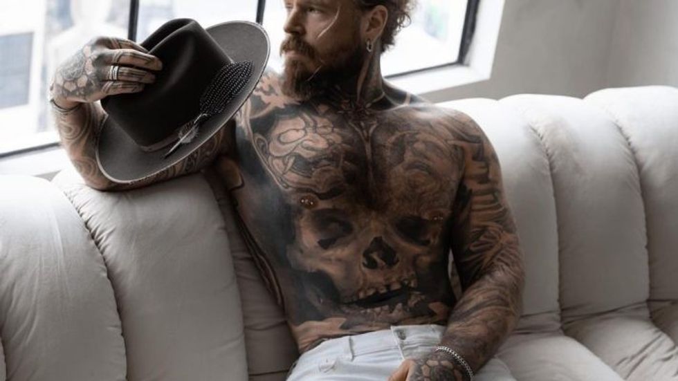 man with tattoos sitting on a couch, holding a hat
