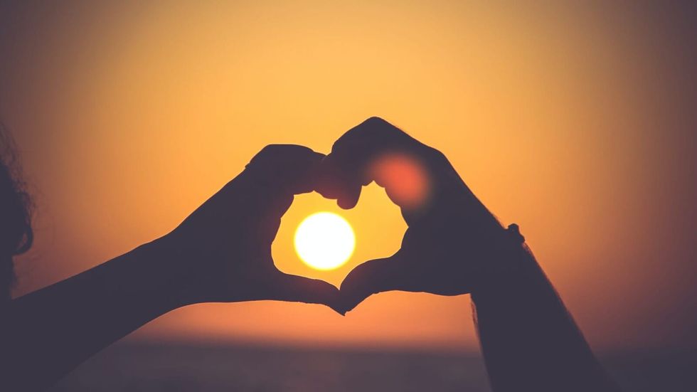 person making a heart with their hands around the setting sun