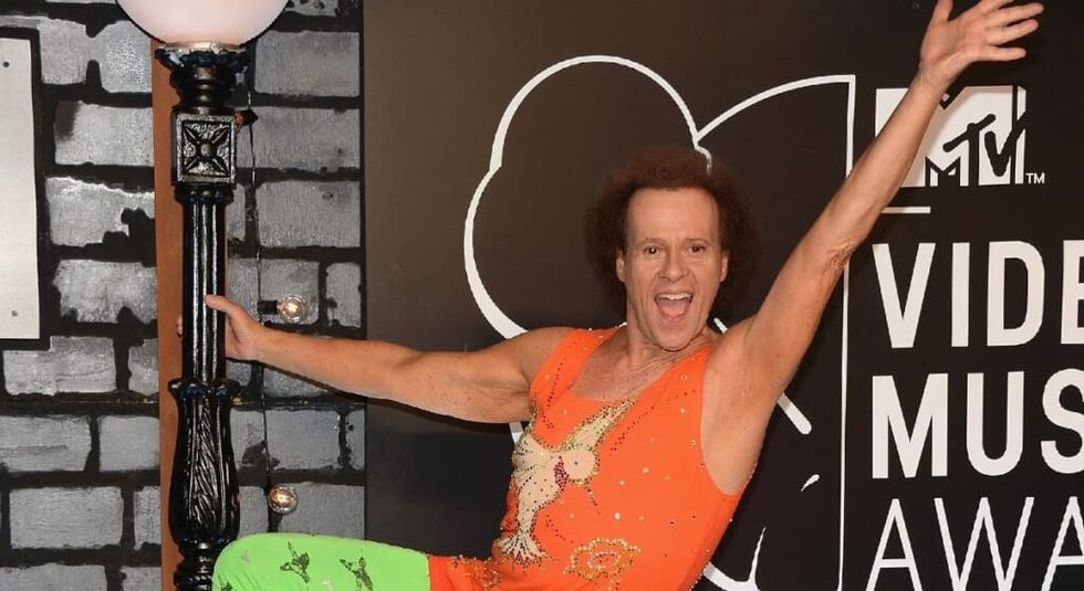 Richard Simmons in orange tank top smiling and raising his arm.