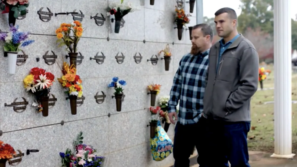 two men standing in front of a wall with bouquets hanging