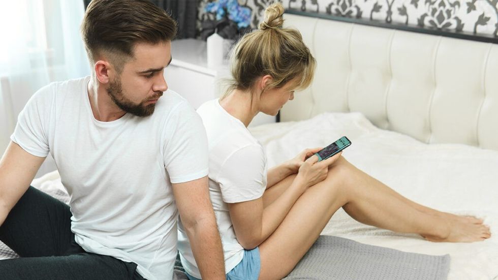 Couple sitting back to back in bed, man looking over shoulder at woman's phone