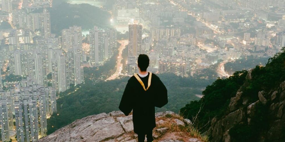 Boy in graduation robe standing on mountaintop overlooking city by Joseph Chan on Unsplash