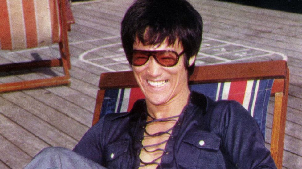 Bruce Lee smiling in 70s clothes sitting in beach chair