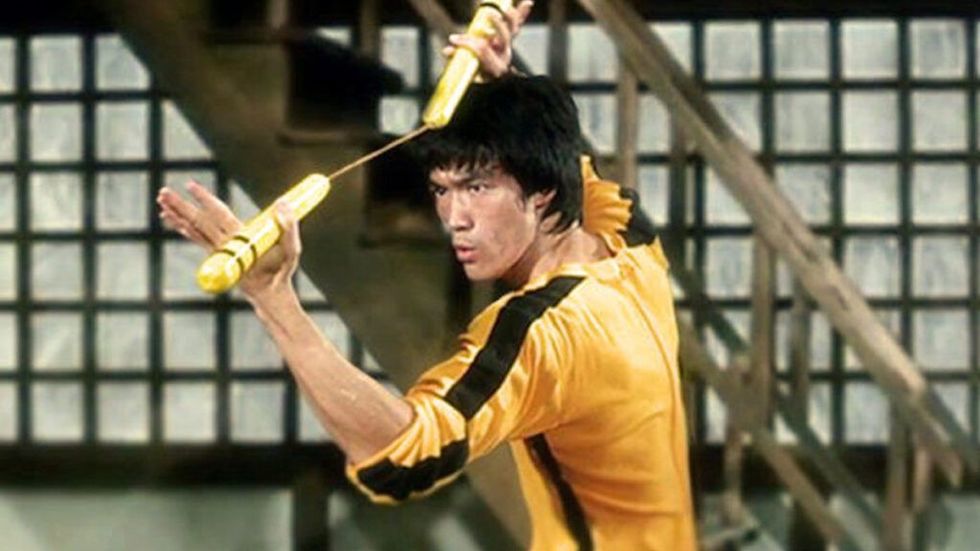 Bruce Lee poartă o salopetă galbenă clasică și ține nunchucks