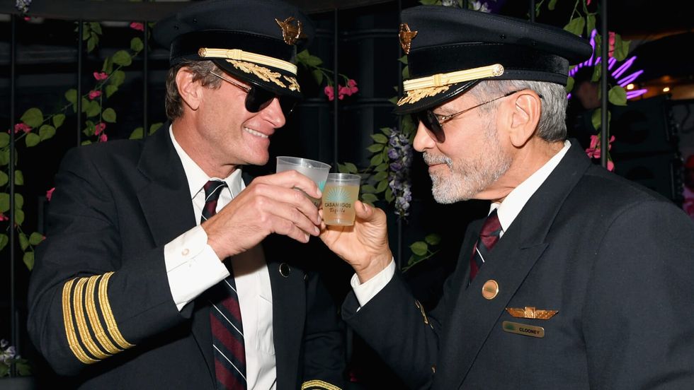 two men wearing black suits and caps clinking drinks together
