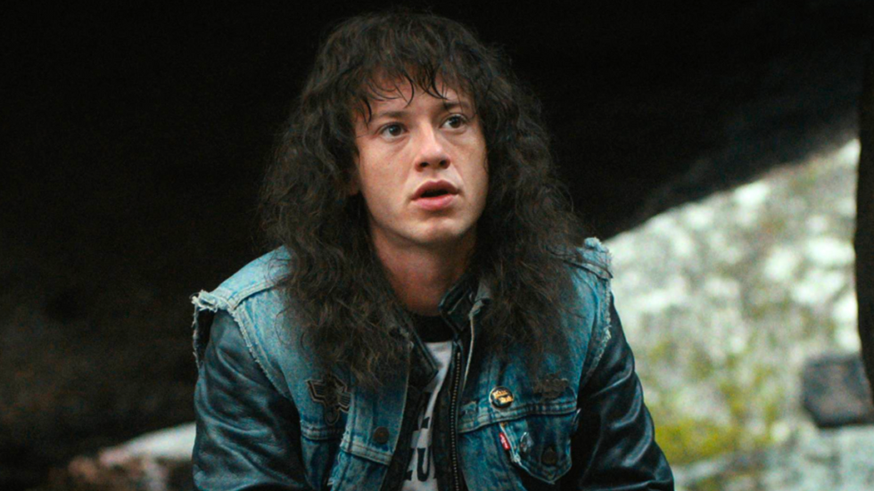 Eddie Munson, a dark-haired man in his early twenties, kneels under a rock. It's a close-up shot as he speaks. He is wearing a leather jacket, a baseball shirt that reads "Hellfire Club," and a cut-out denim vest with pins. His hair is long and shaggy.