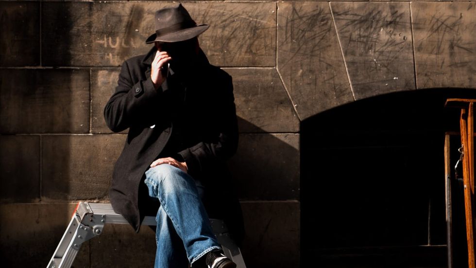 man sitting in the shadows wearing a trench coat and a hat, talking on the phone