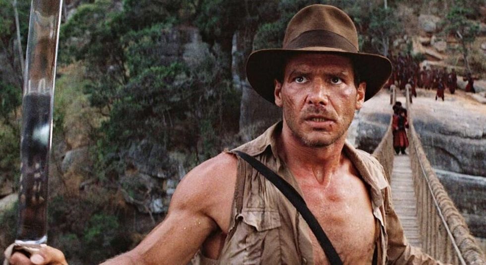 Harrison Ford in Indiana Jones holding a sword in the air while being chased on a bridge