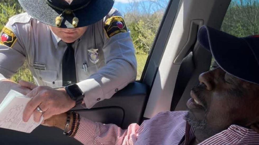 A state trooper is holding a Black man's hand from outside the driver's vehicle