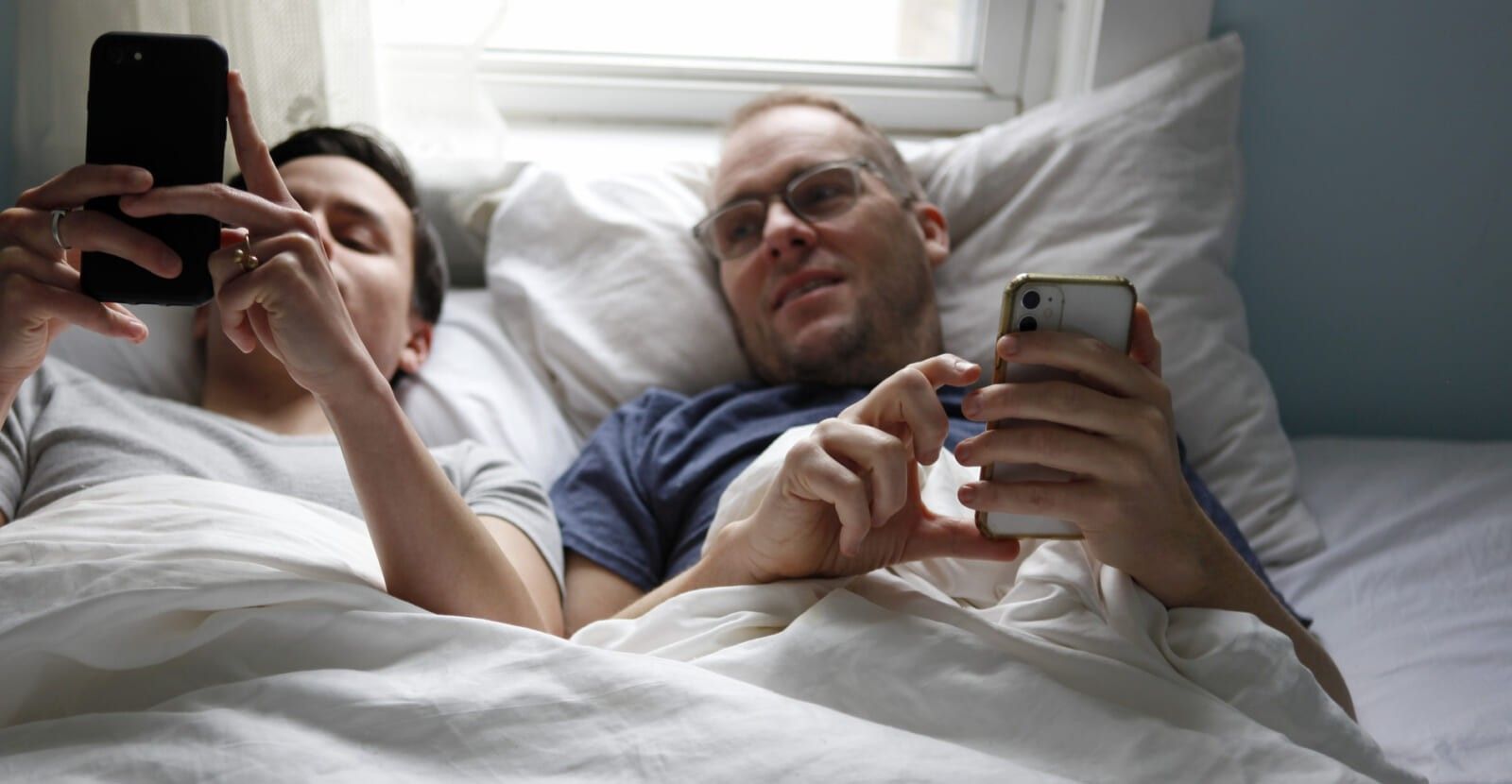 Two men in bed, side by side, looking at mobile phones