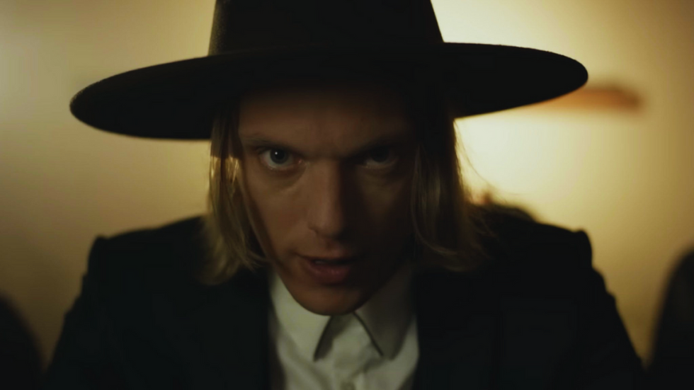 Jamie Campbell Bower wearing a hat, looking like a Southern preacher straight at the camera with an unsettling gaze.