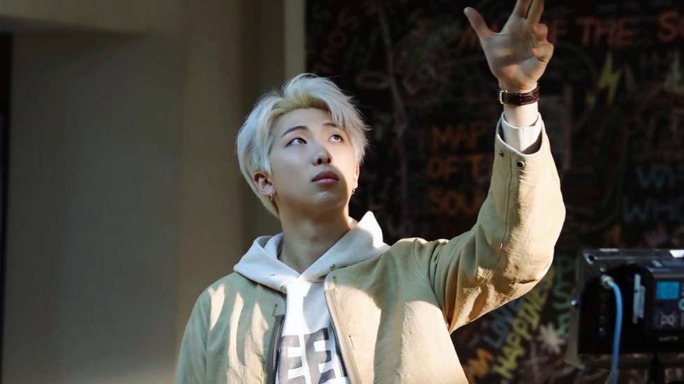 Kim Namjoon, Korean male with blond hair, dressed in a white hoodie and tan jacket, reaches up to a camera out of frame.