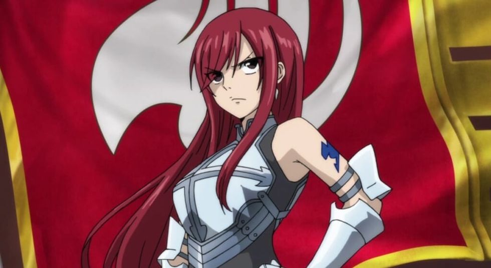 Full body image of Erza Scarlet from Fairy Tail. Erza has a serious expression and has one hand on her hip. She is wearing armor that has a skirt and leaves the shoulders exposed.