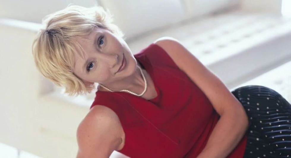 Young Anne Heche dressed in red leaning on a couch.