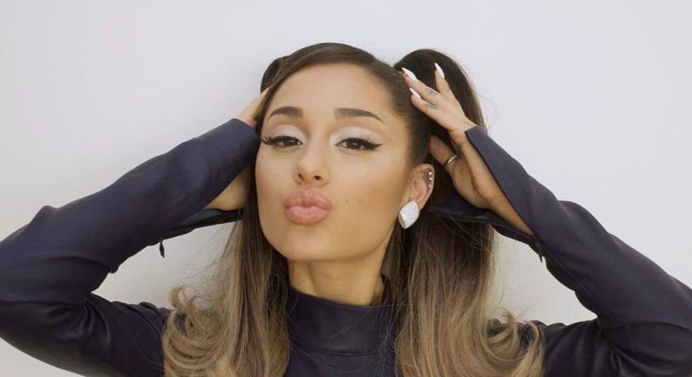 Ariana Grande in a black sweater pulling her hair back and blowing kiss at camera.