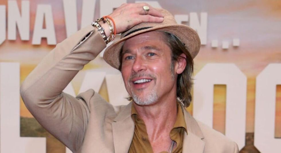 Brad Pitt poartă un costum bej și își pune pălăria în fața camerei.