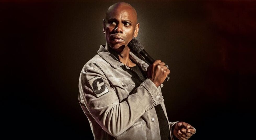 Dave Chappelle wearing green jacket holding a microphone during stand up routine.