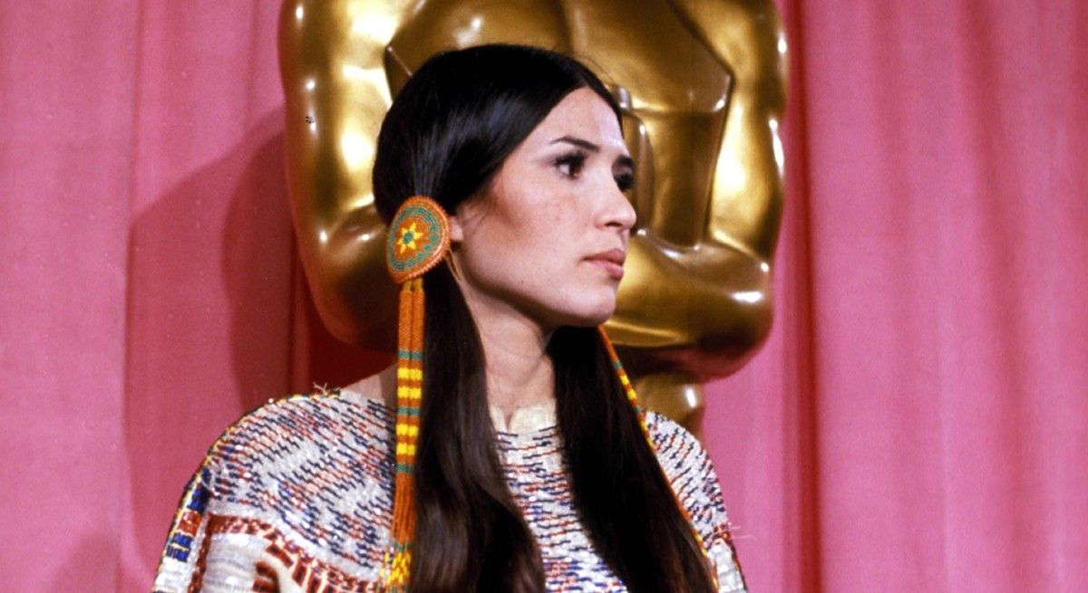 Sacheen Littlefeather standing in front of a large Oscars statue.