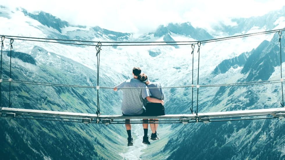 Couple cuddling on a rope bridge looking at a snowy mountain range