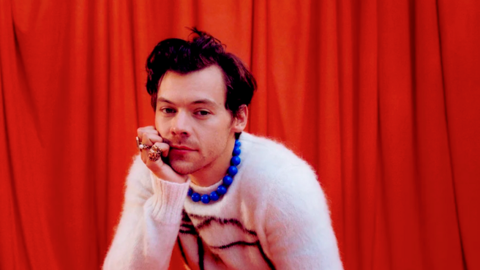 Harry Styles leaning on his palm looking to the camera with an orange curtain in the background.