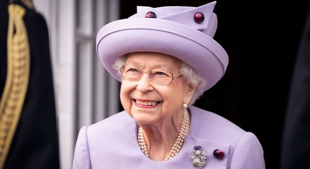 Queen Elizabeth in purple suit and matching purple hat smiling at the camera.