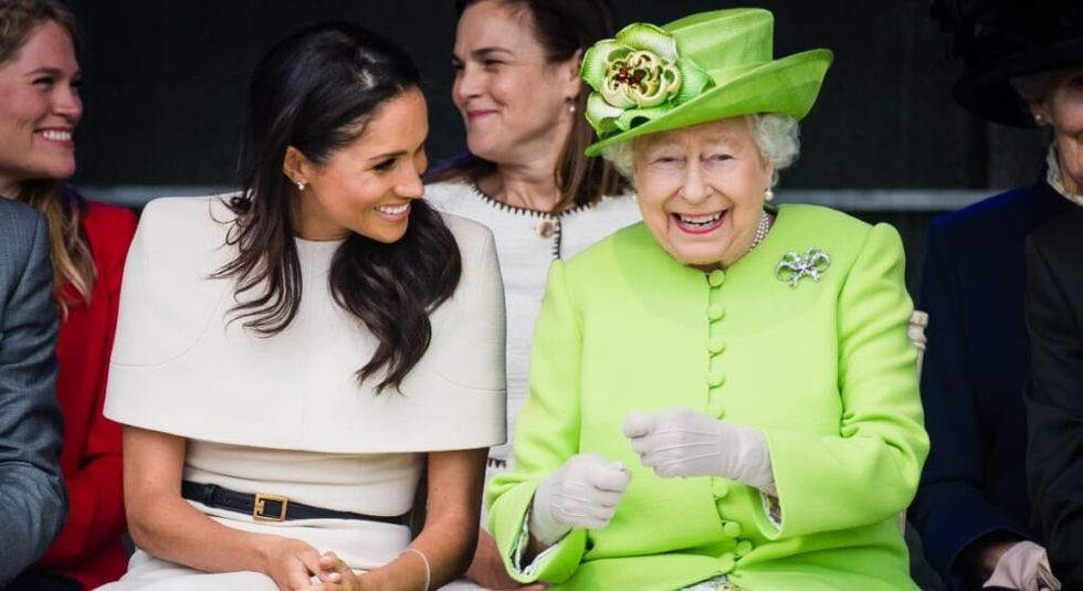 Queen Elizabeth in bright green suit and hat laughing with Meghan Markle who wears a white dress.
