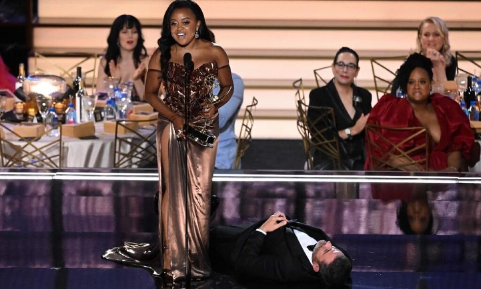 Quinta Brunson accepts her Emmy in a gold dress while Jimmy Kimmel lays on the floor by her feet.