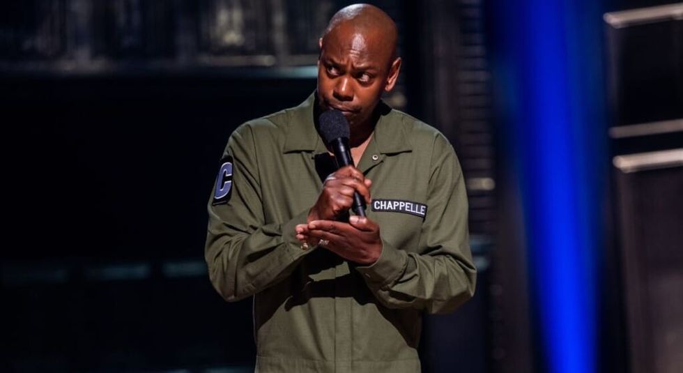 Dave Chappelle in green jumpsuit holding out his hand during a standup routine.