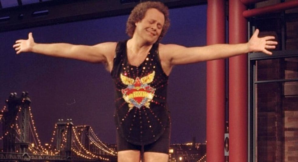 Richard Simmons stands on a desk with his arms outstretched.
