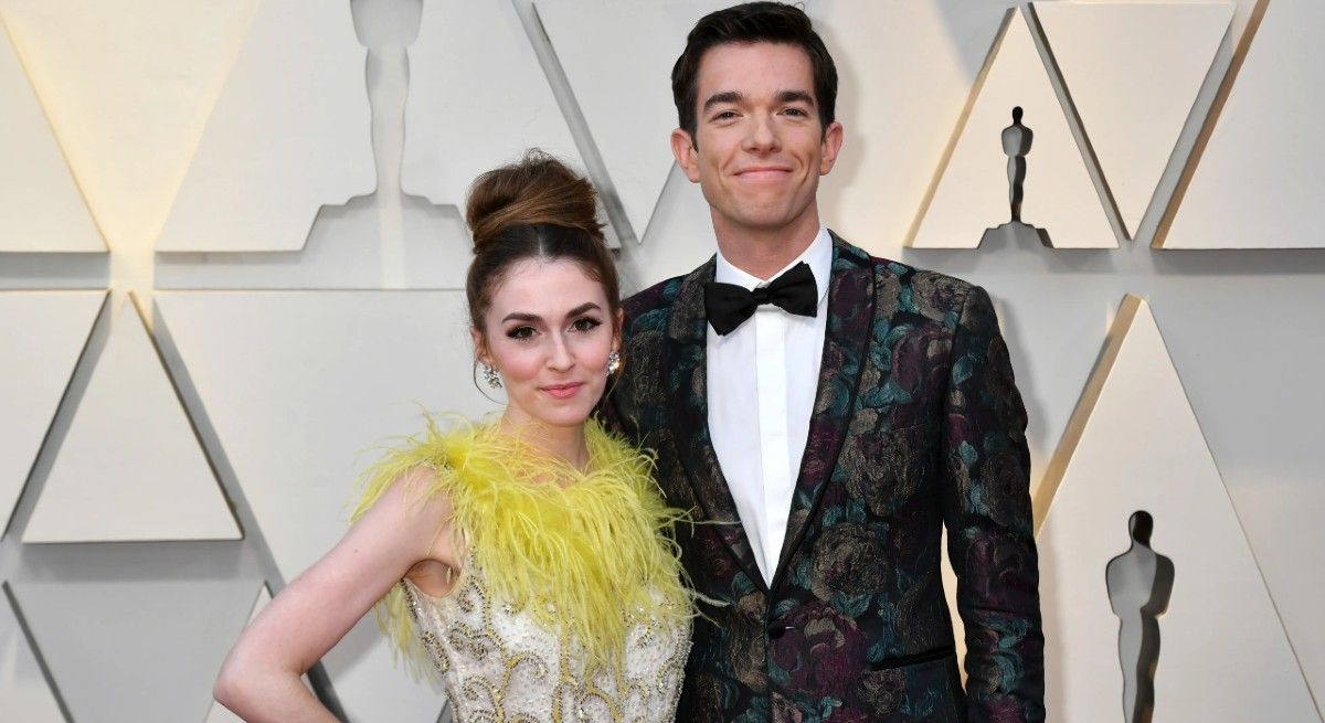 John Mulaney wearing a tuxedo with his arm around his wife who is wearing a yellow dress.