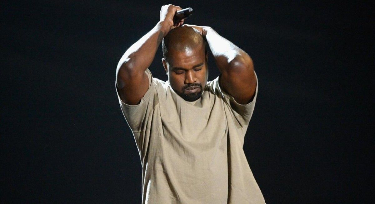 Kanye West in beige shirt holding his head in exasperation.