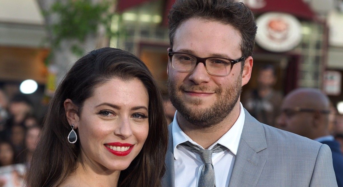 Seth Rogen in grey suit and tie hugging his wife.