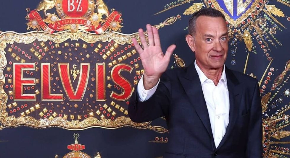 Tom Hanks wearing a dark suit and waving in front of an Elvis sign.