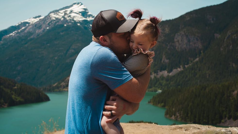 man wearing a blue tshirt kissing a little girl on the cheek in front of mountains