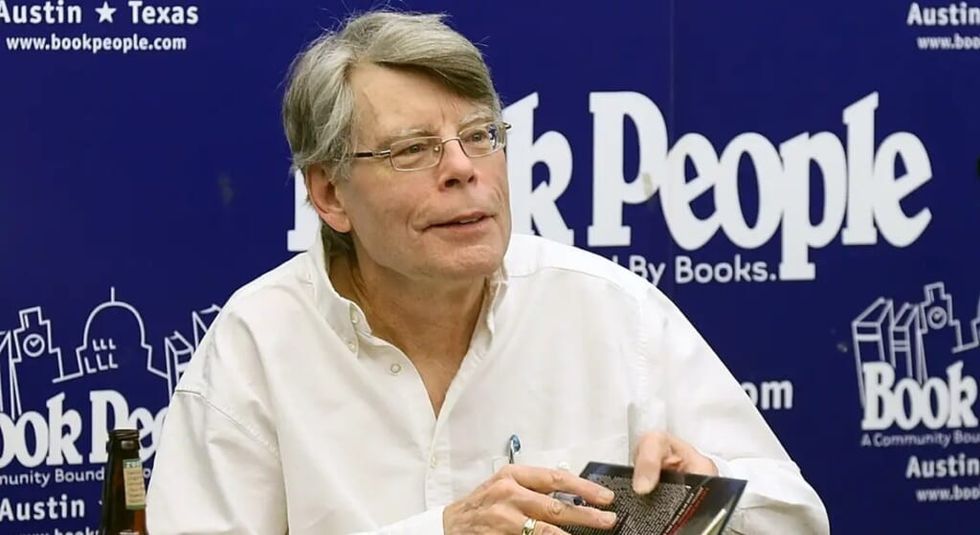 Stephen King white shirt signing books at a desk.