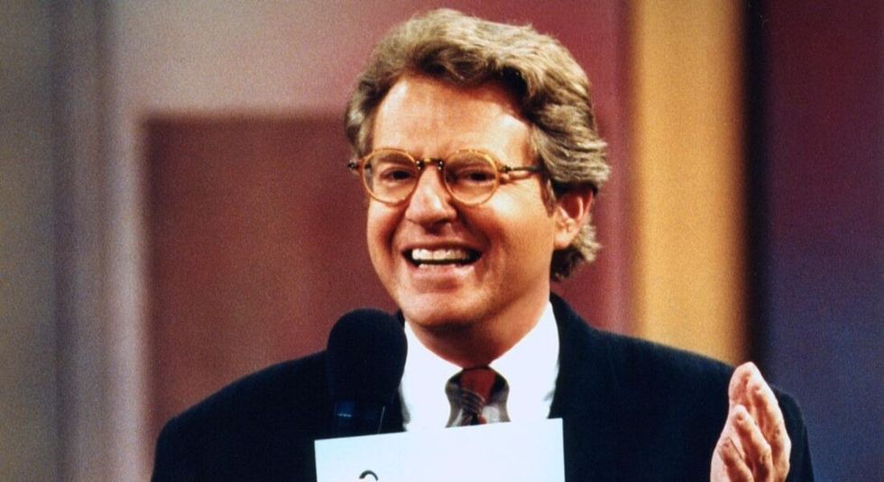 Jerry Springer on the set of his talkshow, smiling and speaking into his microphone.
